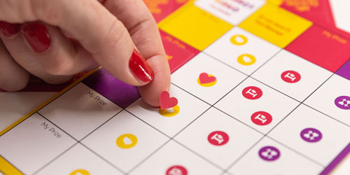 close up of a woman sticking the heart sticker on a bingo sheet of the couple's game - intimacy bingo by joyful couple